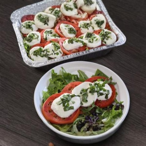 Two servings of Caprese salad with sliced tomatoes, fresh mozzarella, and basil, one in a foil pan and one on a plate.