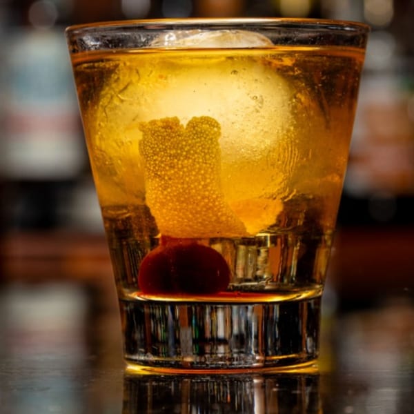 Close-up of an Old Fashioned cocktail with a large ice sphere, orange peel, and cherry.